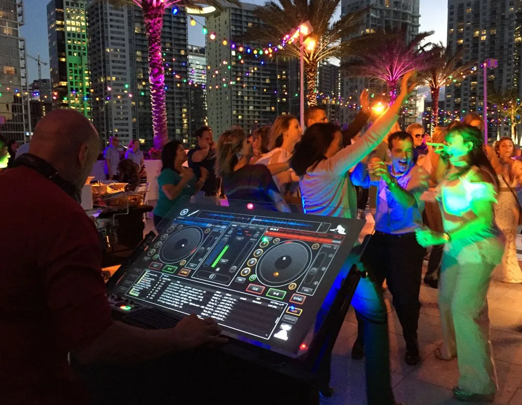 Guests dancing at an evening reception beside a DJ with a lit controller