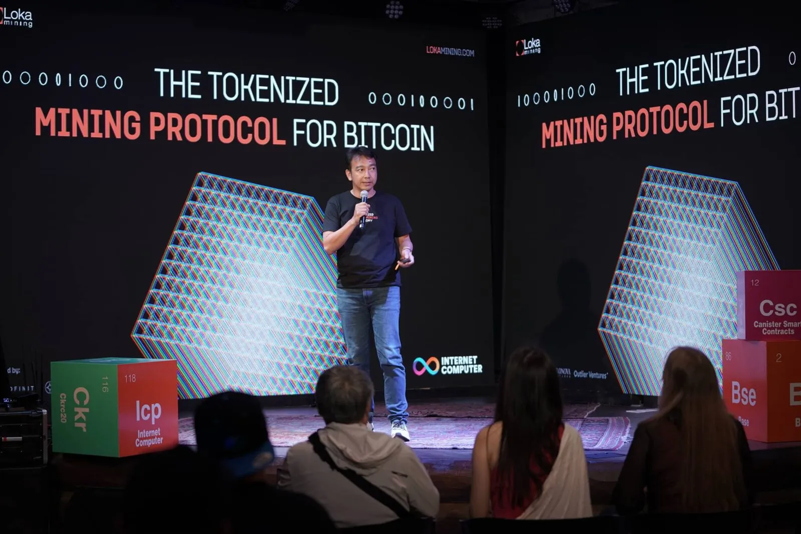 Speaker presenting on a dark conference stage in front of a large projected slide