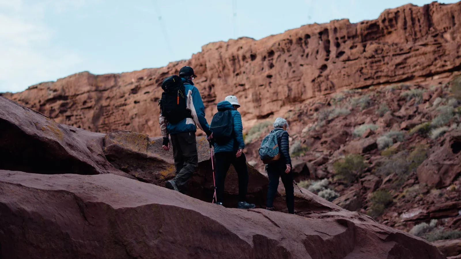 Three hikers with daypacks cross sandstone ledges beneath a red canyon wall on a desert hike during an incentive trip.