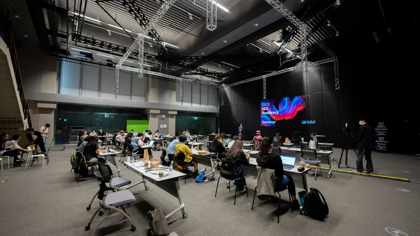 Attendees sit at work tables across a wide, dark event hall facing a large screen with a colorful abstract display.