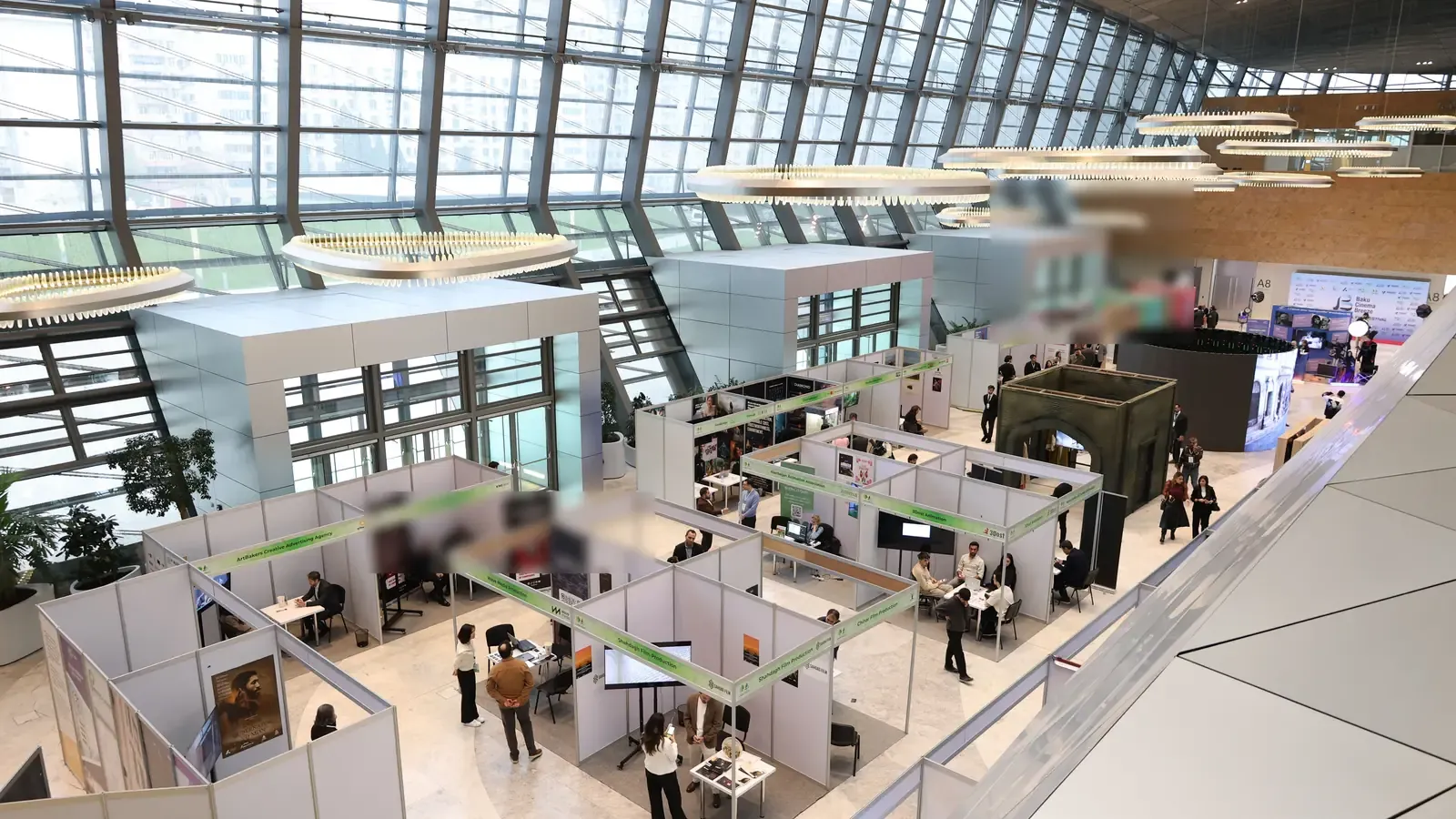 Seen from a balcony, attendees move between rows of white meeting booths on the floor of a bright exhibition hall. A tall glass roof lets daylight pour over the space.