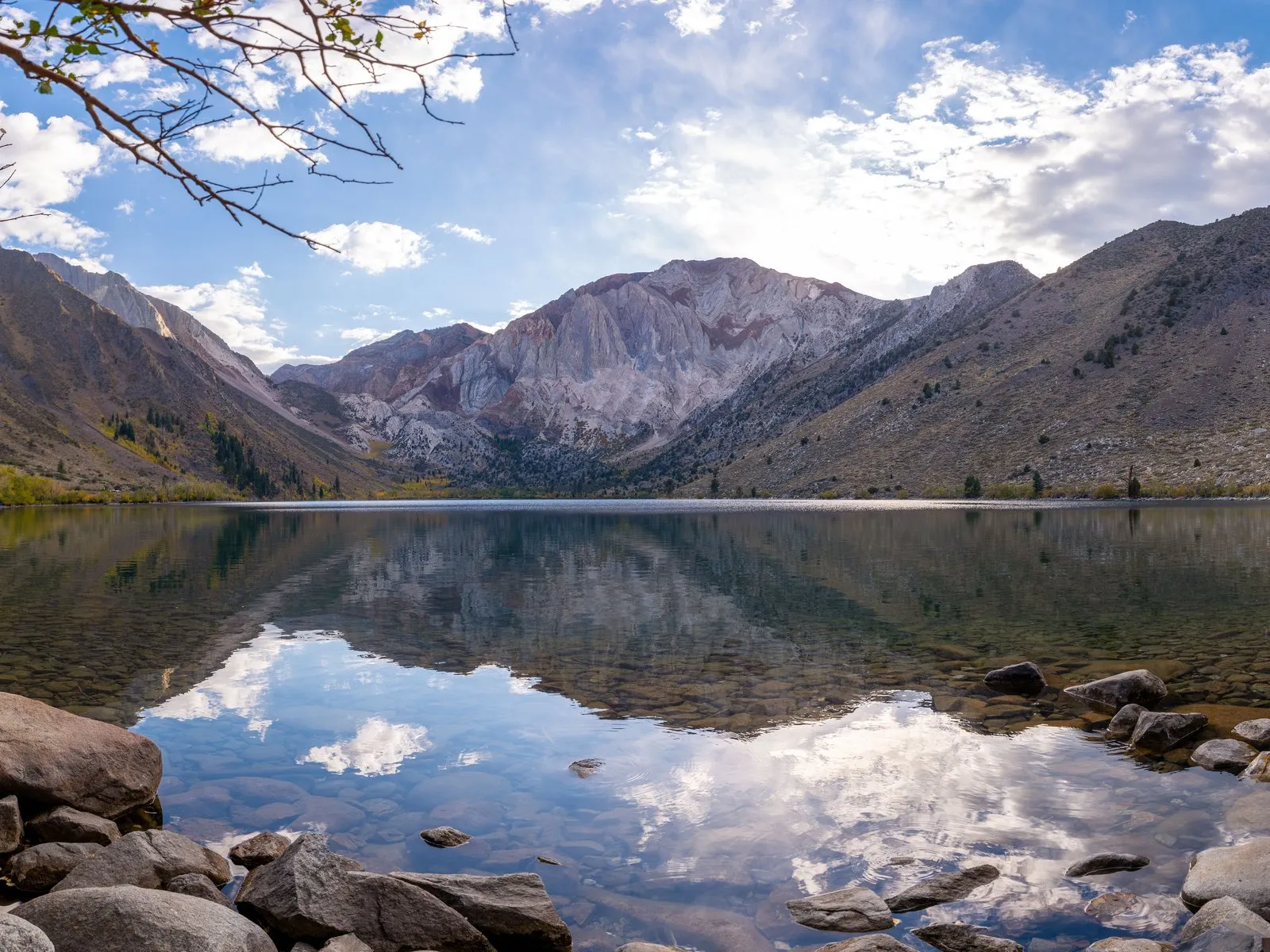 Convict Lake lies still beneath banded, bare peaks just outside Mammoth Lakes, with early-fall aspens turning gold along the far shore. Clouds and mountains reflect in the clear water above a shallow, rocky bottom.