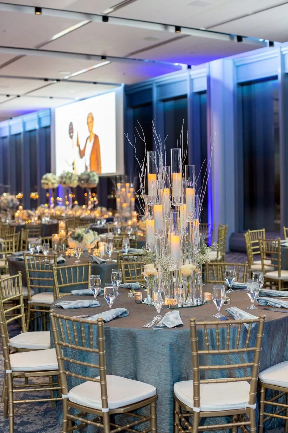 Round dinner tables with candle centerpieces and projection screens at a corporate event