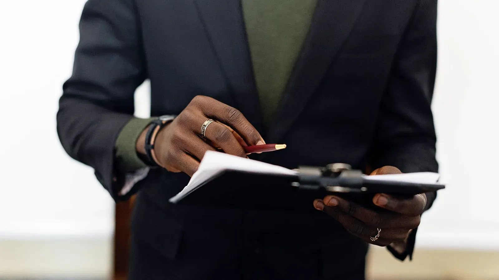 A person in a dark blazer holds a pen and a clipboard with papers, seen from the chest down against a bright blurred background.
