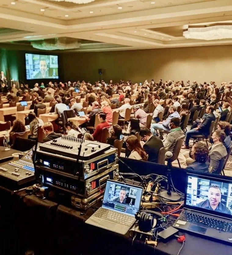 Seated audience in a large general session room with a production desk and screens