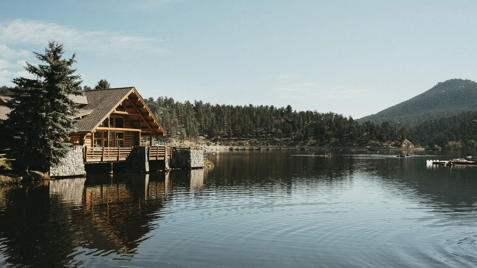 A timber lodge with a wraparound deck sits on a calm mountain lake, with pine forest and a wooded ridge behind it.