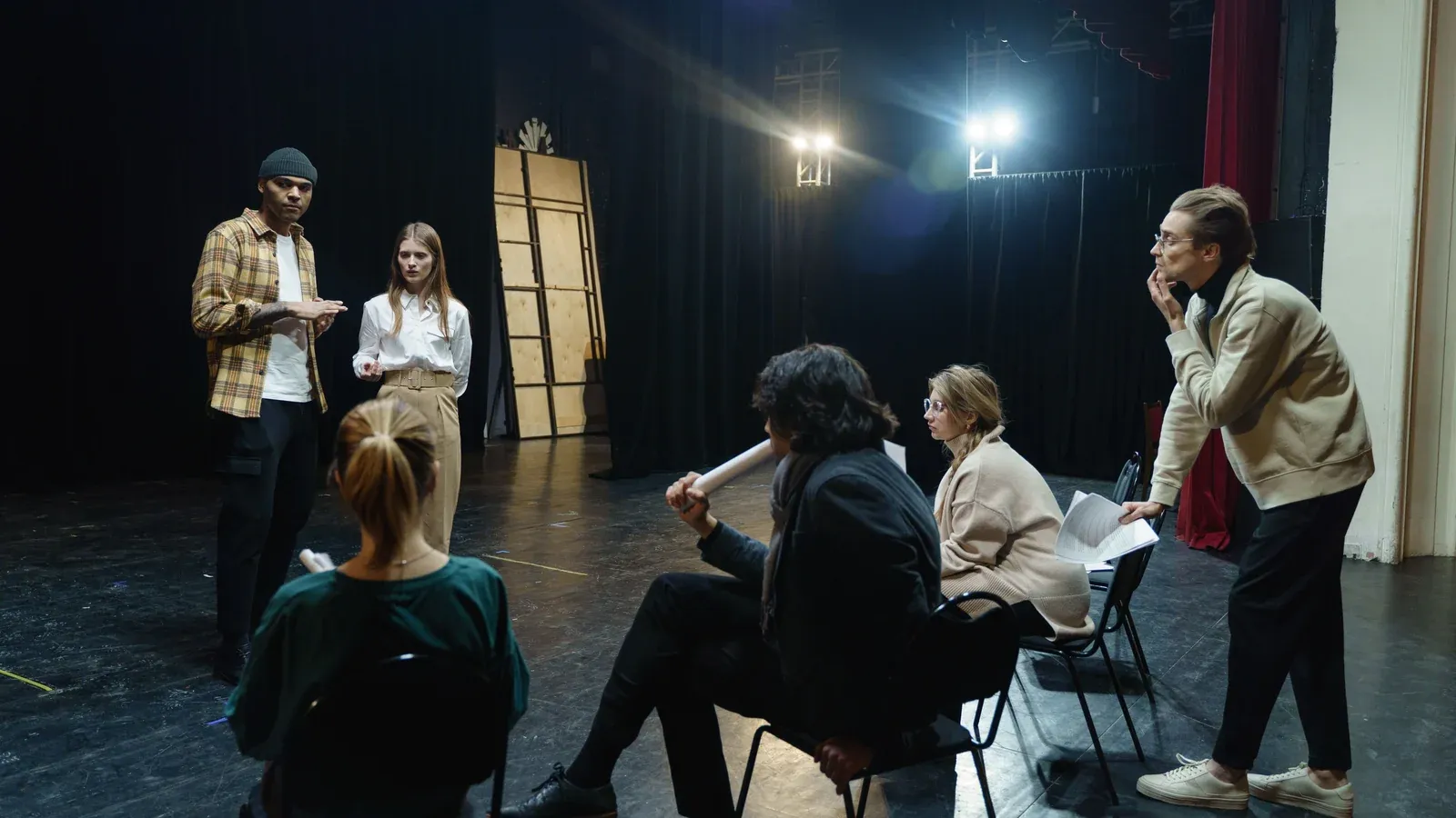 Six adults gather on a dimly lit theater stage, three seated and three standing, some holding papers.