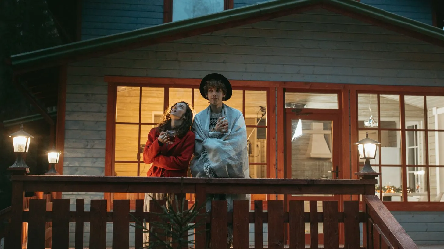 Two people stand on the lit porch of a wooden cabin among tall pines at dusk during a weekend company retreat.
