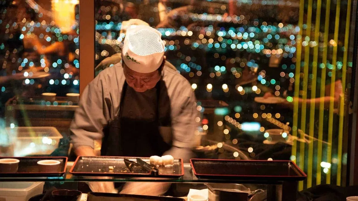 Chef preparing sushi at a counter with city lights behind