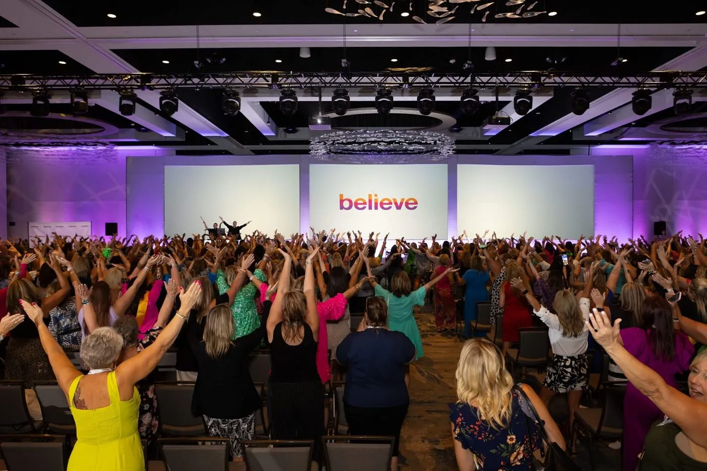 Conference attendees with raised arms in front of a stage screen reading believe