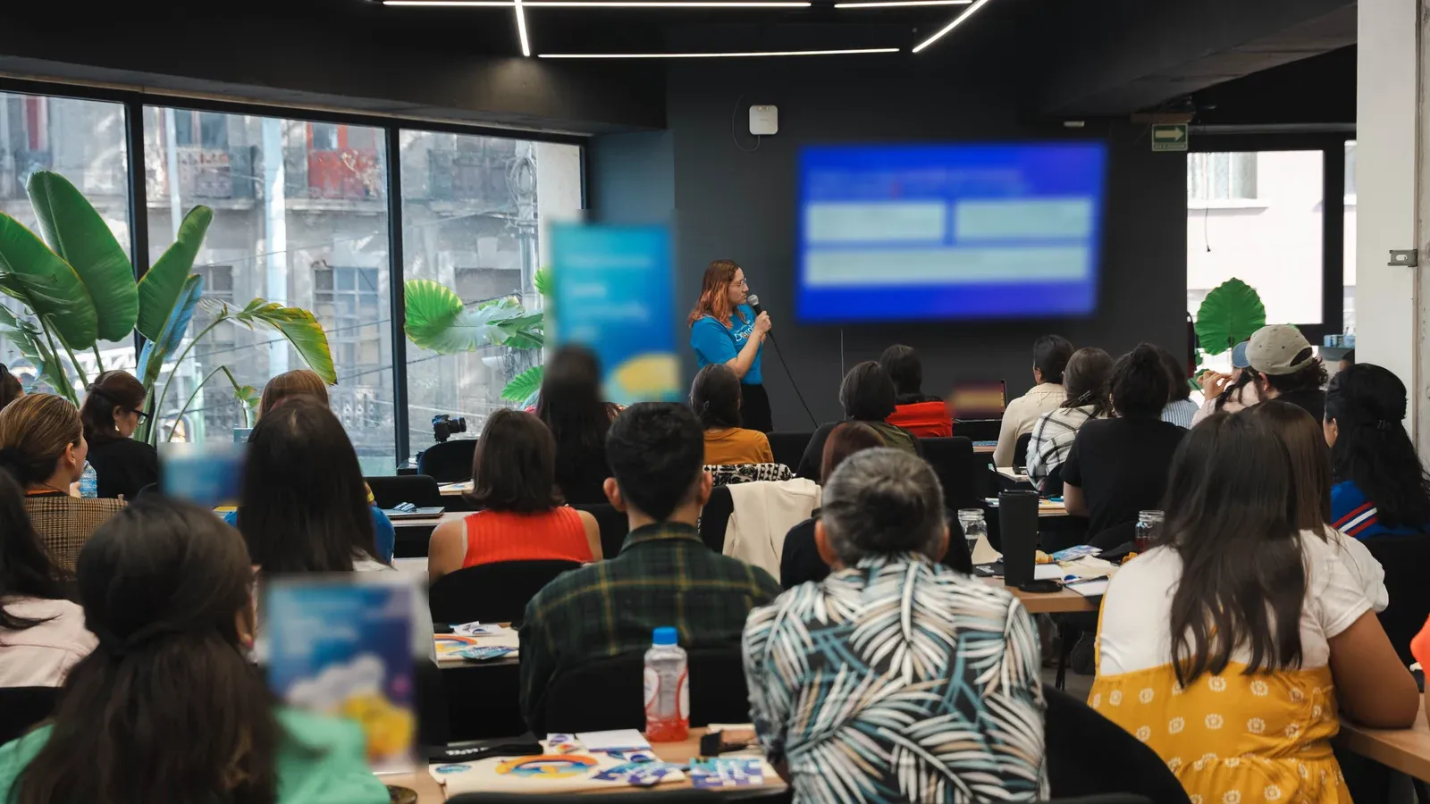 A presenter with a microphone addresses a packed room of attendees seated at long tables. Tall windows and leafy plants frame the space beside a large screen.