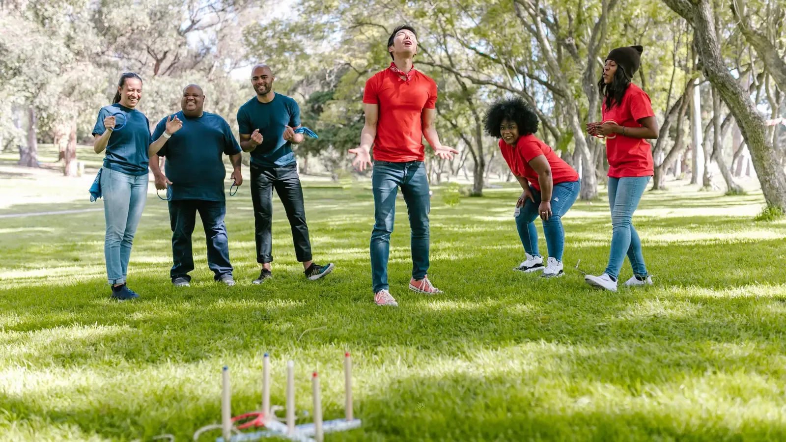 Six people in matching teal and red T-shirts laugh and react while playing ring toss on a sunny, tree-shaded park lawn.
