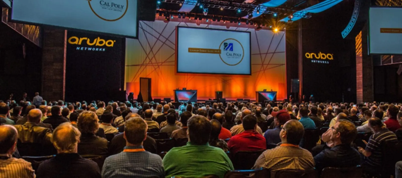 Audience at an Aruba general session with stage screens and orange lighting