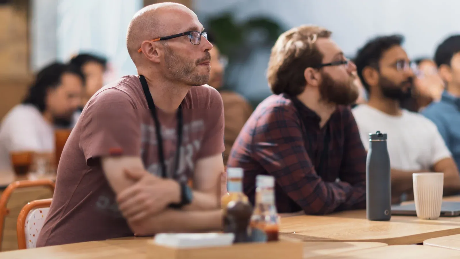 Attendees sit along a row of wooden tables, listening closely to a session at the front of the room. A man in glasses leans forward with folded arms beside a colleague in a plaid shirt.