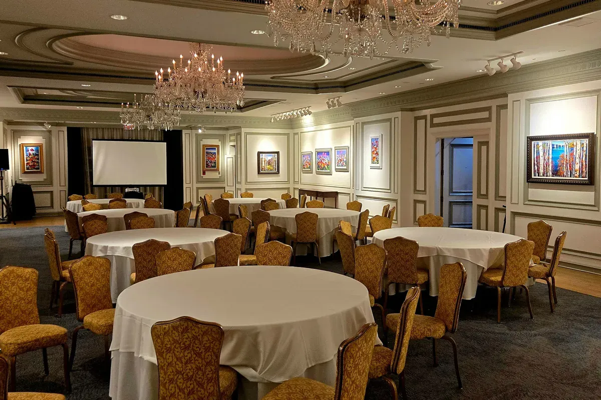 Round tables draped in white linen and ringed by gold upholstered chairs fill a hotel function room under crystal chandeliers. A blank projection screen stands at the far end, and framed paintings hang on the paneled walls.