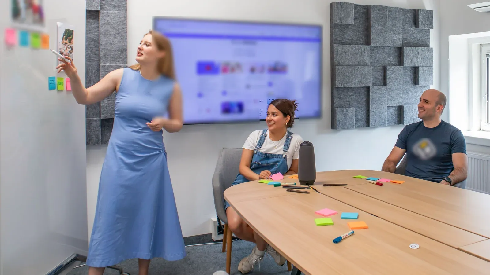 A woman in a blue dress points to sticky notes on a wall as she leads a small breakout session. Two colleagues at a curved wooden table smile and follow her plan.