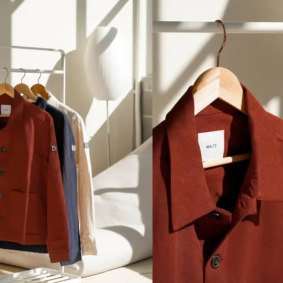 Rust, navy and cream cotton work jackets hang on wooden hangers on a rail, beside a close-up of the rust jacket's point collar, woven neck label and dark buttons
