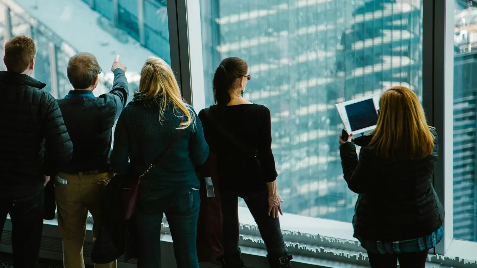 A group of five people seen from behind look out of a tall window over glass high-rise buildings, one holding a tablet.