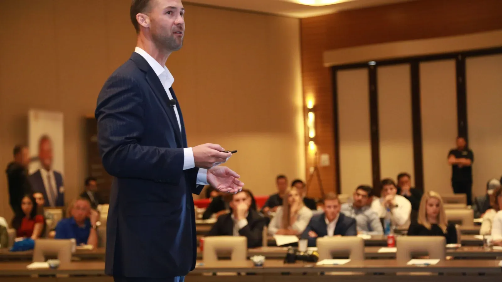 A presenter in a navy suit speaks to a full conference room of seated attendees. Rows of tables with laptops face him under warm hall lighting.
