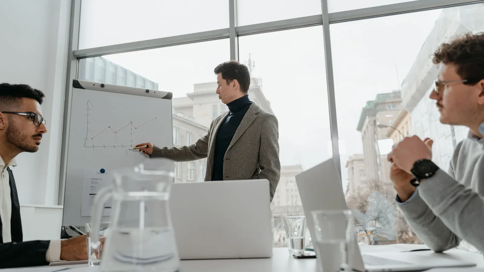 A presenter points to a chart on a whiteboard while colleagues listen at a table. Tall windows fill the meeting room with daylight.