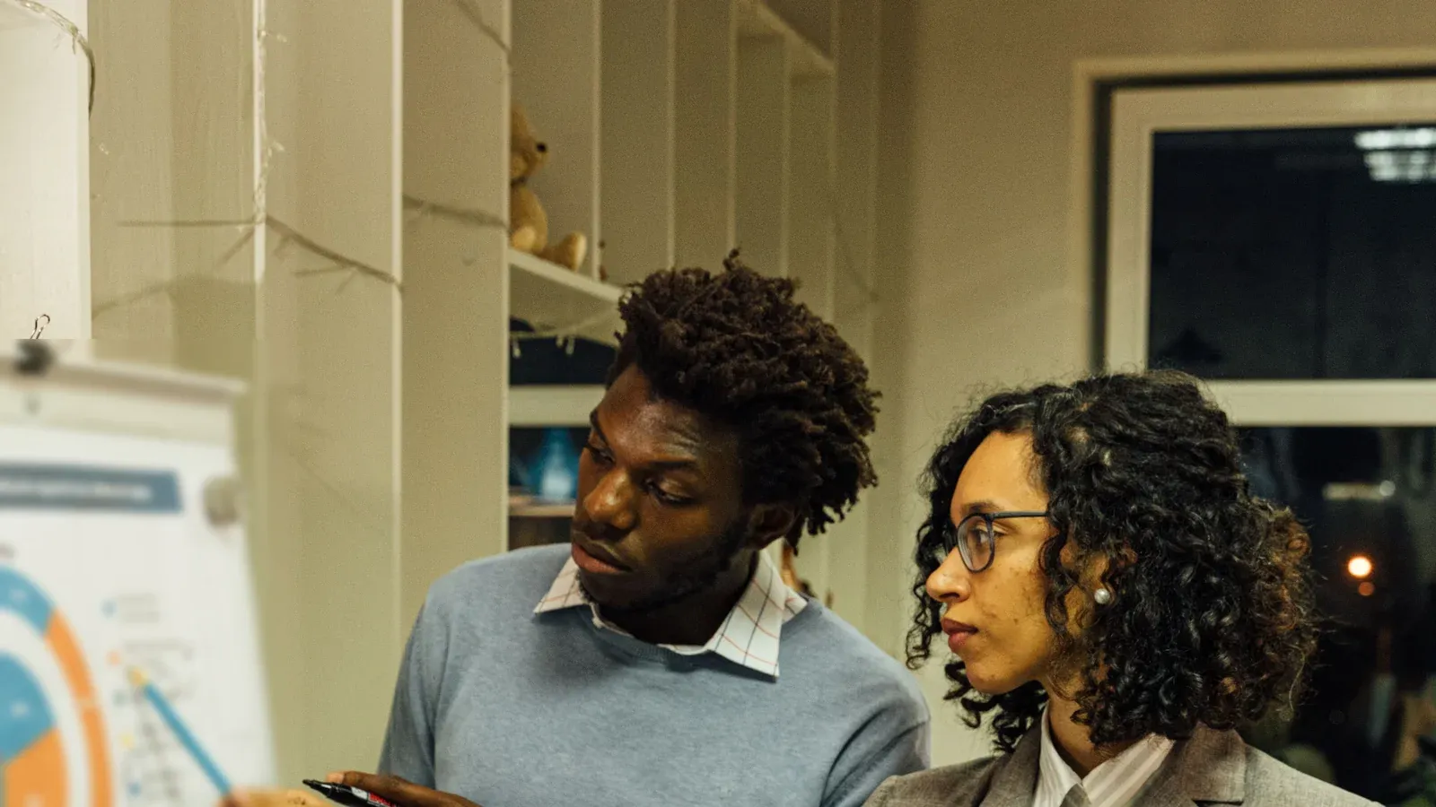 Two colleagues talk through results beside a flip chart in a warm office. Both look toward the chart as they discuss the next steps.