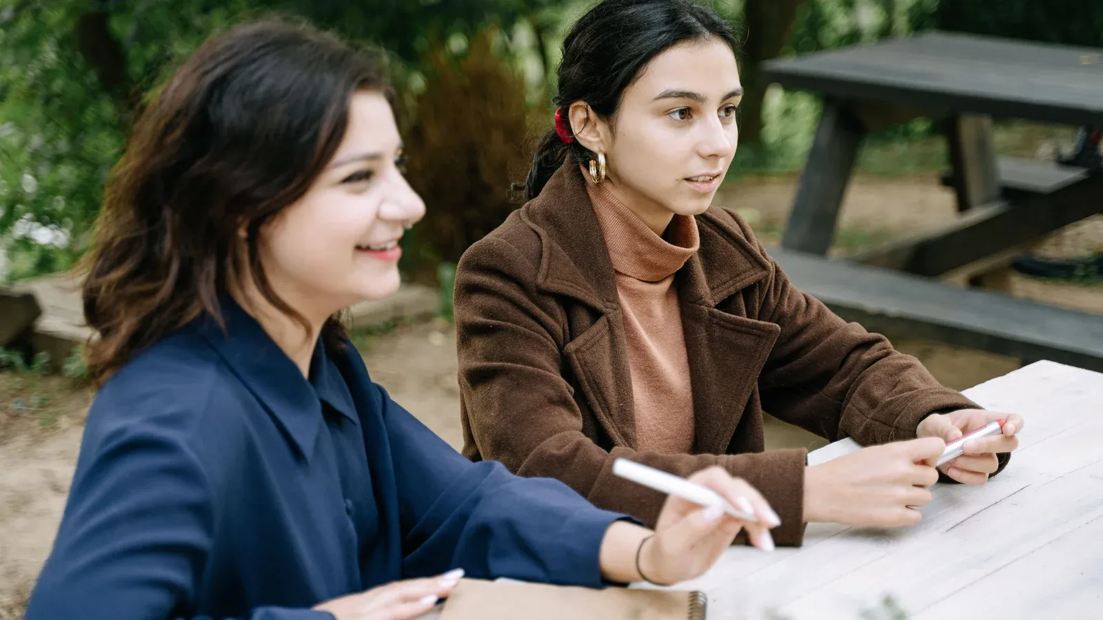 Two colleagues sit side by side at an outdoor wooden table, pens in hand, during a planning session. Green trees and an empty picnic bench fill the background.