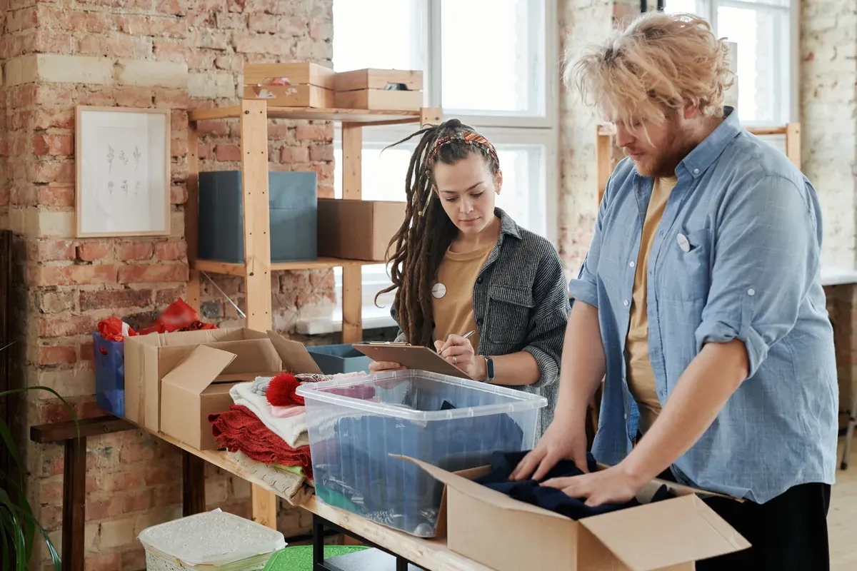 Two volunteers sort donated clothing into boxes and a clear crate at a long wooden table, one checking items off on a clipboard. Packing donations together is an easy way to turn an end-of-year gathering toward giving.