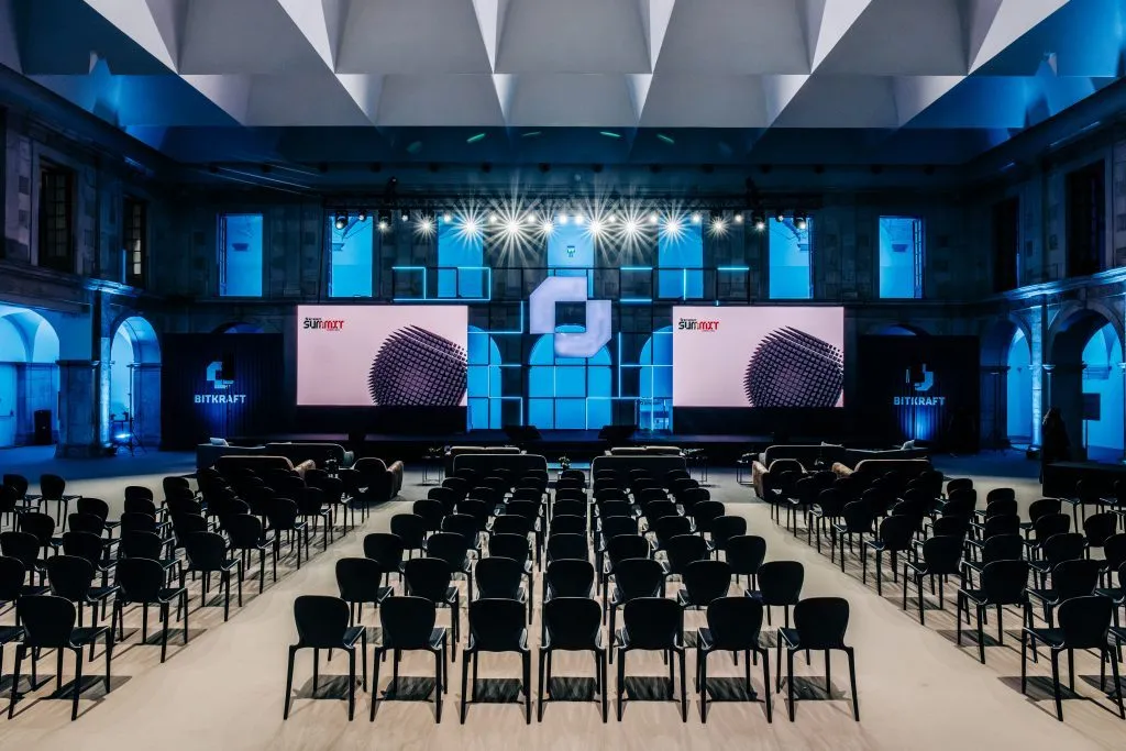 Conference hall with rows of chairs facing a lit stage and screens