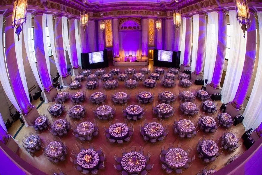 Historic ballroom with round tables lit in purple and screens on both sides of the stage