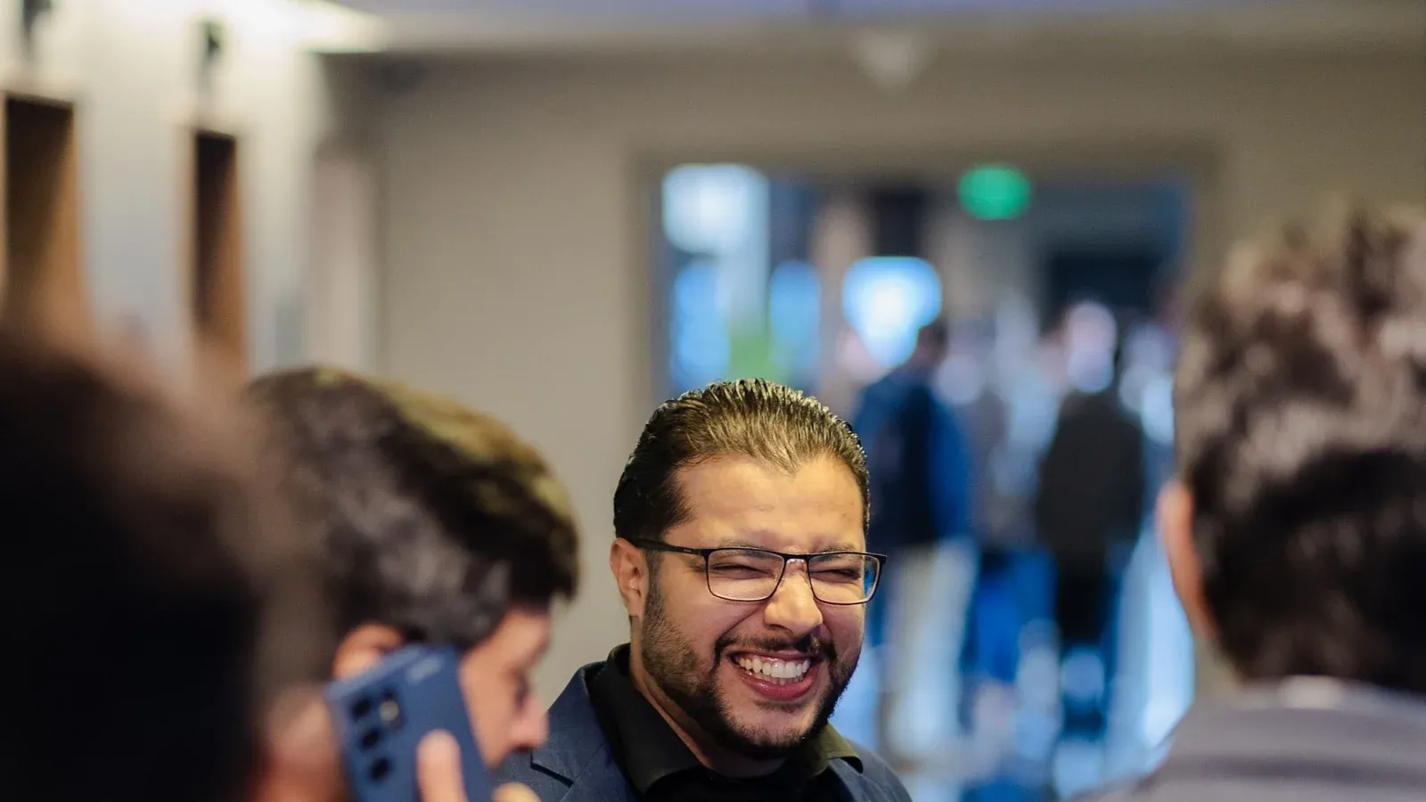 Professionals chat in small groups during an indoor networking session. A man in glasses laughs as he talks with colleagues.