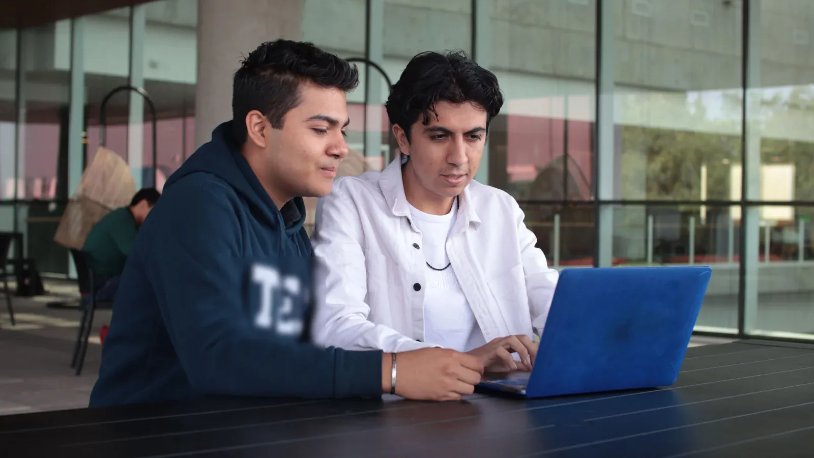 Two colleagues work side by side on a laptop at a long table. Tall glass windows behind them let in soft daylight.