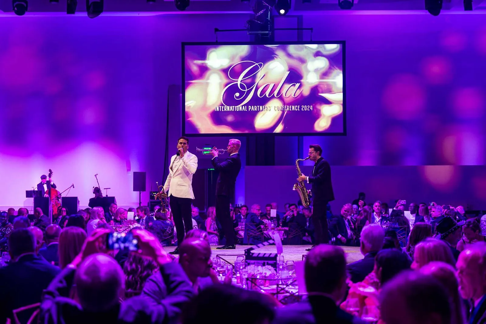 Presenters on stage with a purple lit screen in front of a seated audience