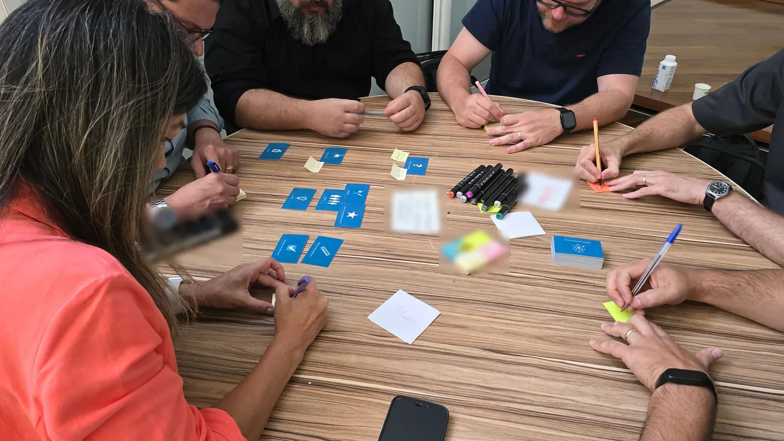 A breakout group gathers around a round wooden table covered with blue cards, sticky notes and markers. Several people write at once as they work through the exercise together.