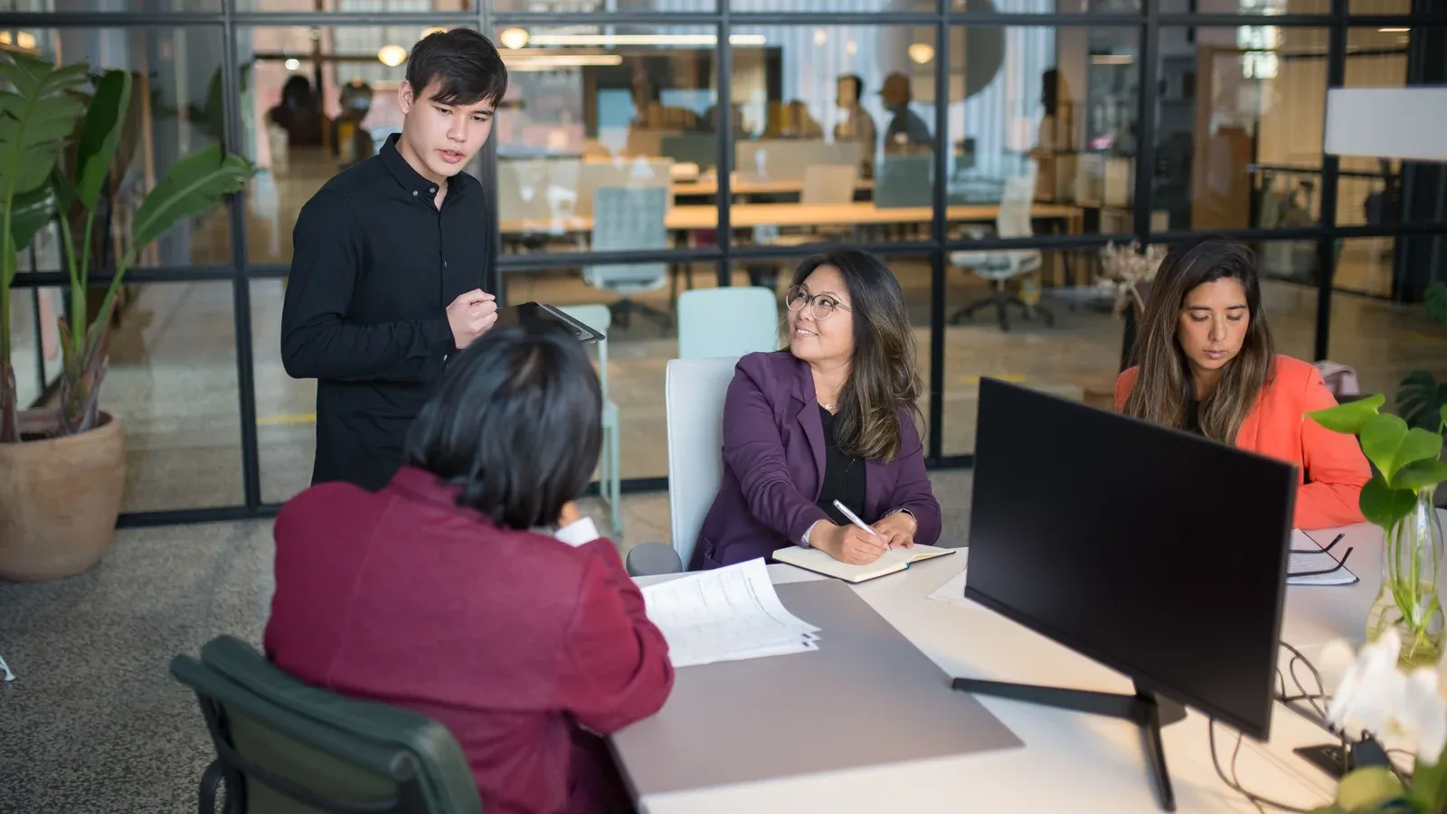 A small team meets around a desk with papers and a monitor in a glass-walled open office during a company offsite.