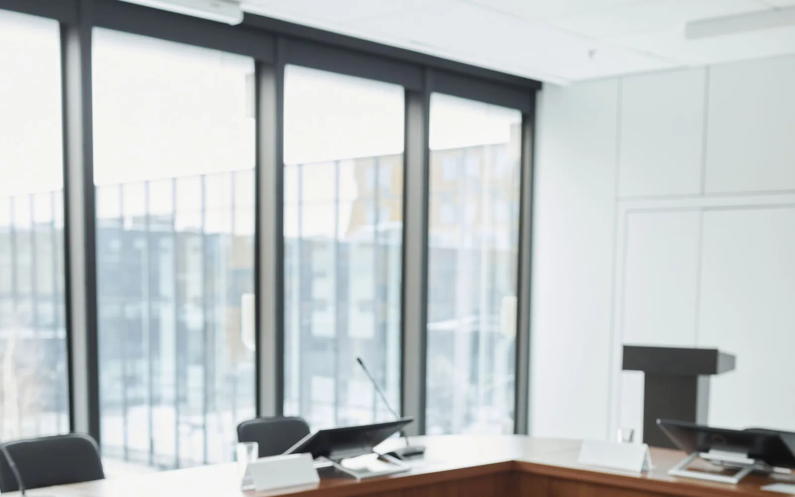 A conference room with microphones, screens, and a U-shaped table beside tall windows.
